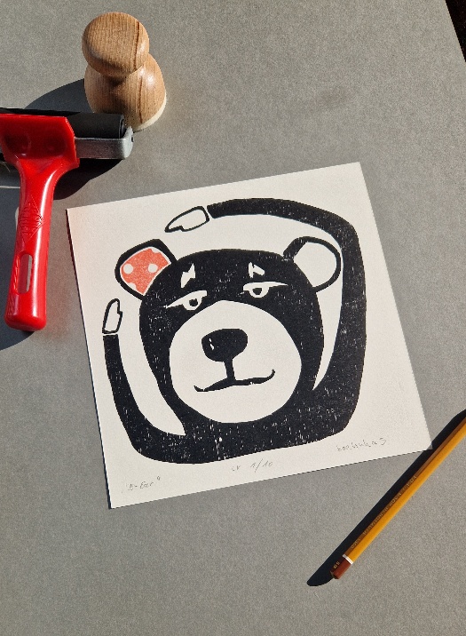 Schwarzer Comicbär,der mit beiden Händen auf sein rechtes, rotes Ohr deutet, gedruckt auf weißem Papier