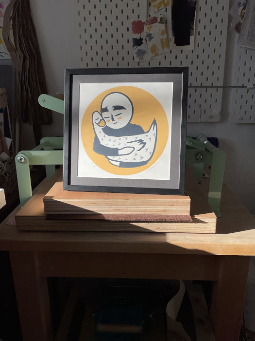 Stilisierte Comicfigur ohne Haare und geschlossenen Augen, eine Gans haltend, mit gelbem Hintergrund gedruckt auf weißem Papier